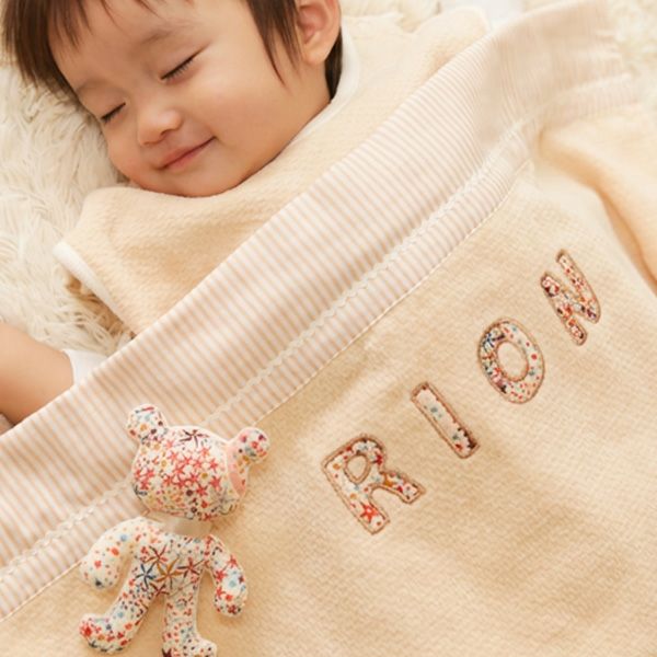 お名前入りの刺繍が施されたクリーム色のタオルケットと花柄のにぎにぎと眠る赤ちゃん